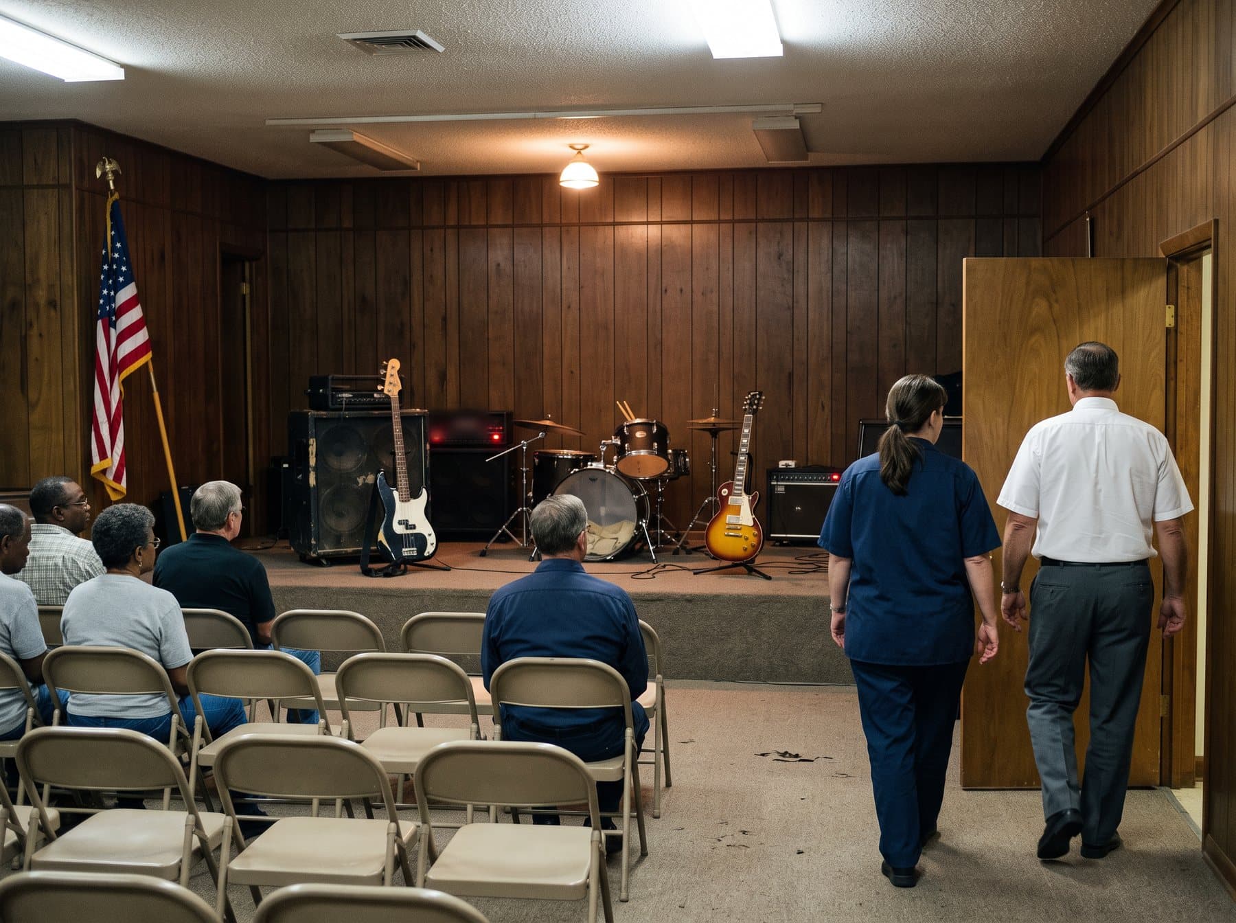 A veterans' post hall with wood paneling and folding chairs, a band leaving a low stage with amplifiers still humming